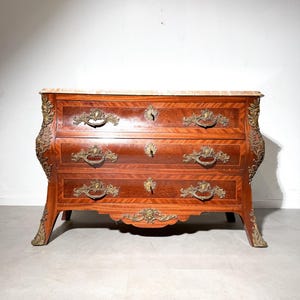 Puede incluir: Cómoda de madera antigua con tres cajones y herrajes de latón ornamentados. La cómoda tiene una tapa de mármol y está hecha de madera de color marrón rojizo. Las patas son curvas y el diseño es antiguo.