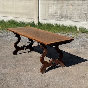 May include: A rectangular wooden table with a smooth, medium-brown surface. The table features two ornate, curved trestle legs connected by a thin, dark metal support rod that runs horizontally between the two base structures.