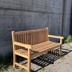 May include: A light brown wooden bench featuring a slatted backrest, a slatted seat, and two armrests. The bench is constructed with vertical slats on the back and horizontal slats on the seat, supported by four sturdy legs and a frame structure.