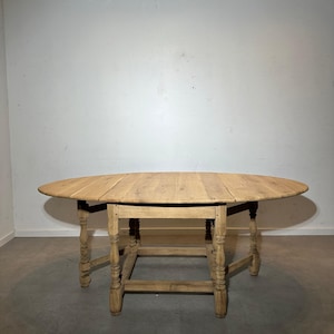 Puede incluir: Una mesa de madera clara de forma ovalada con hojas abatibles. La mesa tiene un aspecto envejecido con la veta de la madera visible. Las patas están intrincadamente talladas y la mesa está contra una pared blanca lisa.