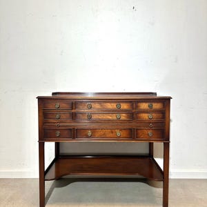 May include: A dark brown wooden desk with multiple drawers and brass hardware. The desk has a rectangular top and a lower shelf. The drawers are arranged in a grid pattern, and the desk is supported by four legs.