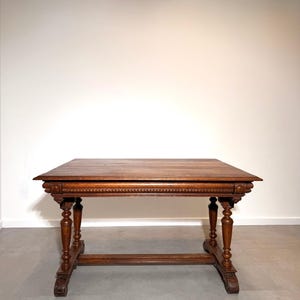 May include: A rectangular wooden table with a dark brown finish, featuring a flat top surface and a decorative carved apron. The table is supported by four turned legs connected by a horizontal stretcher bar, with carved feet at the base.