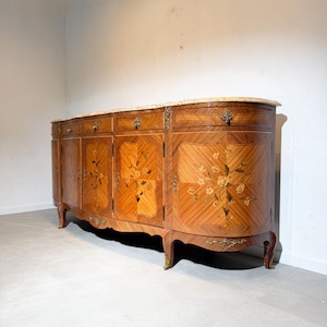 Puede incluir: Aparador antiguo de madera con tapa de mármol. El mueble presenta intrincados patrones de madera incrustada y diseños florales en las puertas. Tiene herrajes de latón y patas curvas, mostrando un estilo clásico y elegante.