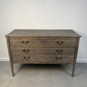 May include: A weathered, wooden dresser with three drawers and ornate dark metal handles. The dresser has a rectangular top and sits on four slender, tapered legs. The wood has a natural, gray-brown finish, giving it a rustic appearance.
