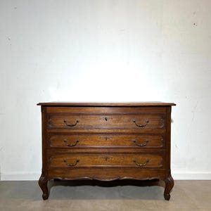 Puede incluir: Cómoda de madera de tres cajones con acabado marrón oscuro. El mueble tiene tiradores de latón ornamentados y detalles decorativos. Las patas son curvas y el estilo general es antiguo.