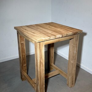 Könnte beinhalten: Ein hoher, quadratischer Stehtisch aus Holz mit einer natürlichen, hellbraunen Holzmaserung. Der Tisch besteht aus vier stabilen vertikalen Beinen, die durch horizontale Stützstreben nahe der Basis verbunden sind und eine flache Tischplatte tragen.
