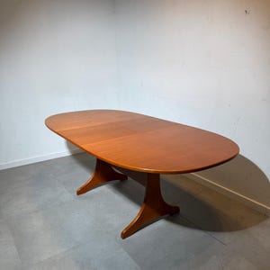 May include: An oval-shaped wooden dining table with a smooth, warm-toned finish. The table features a central pedestal base with two wide, curved feet that extend outwards for stability, supporting the expansive, flat tabletop surface.