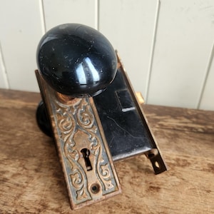 May include: Antique door hardware with a black ceramic knob and ornate brass trim. The door hardware has a keyhole and a decorative design.