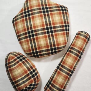 May include: Three plaid fabric items in shades of brown, tan, and black. The items include a mitten-shaped pot holder with a hanging loop, an oval-shaped item, and a long, cylindrical item. The items are arranged on a white surface.