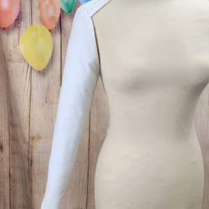 May include: White long-sleeved garment with a single sleeve, worn on a white mannequin. The garment is made of a smooth, white fabric. The mannequin is standing on a wooden surface with a colorful background.