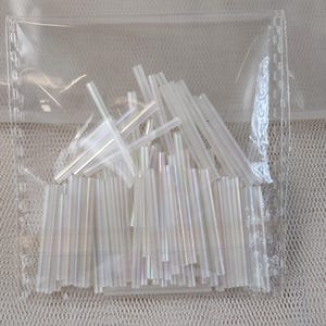 May include: A clear plastic bag filled with iridescent glass beads. The beads are long and thin, and they have a shimmery finish.