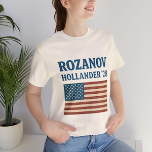 May include: Cream-colored t-shirt featuring the text "ROZANOV HOLLANDER '28" in blue above a vintage American flag graphic. The flag has a blue square with white stars and red and white stripes.