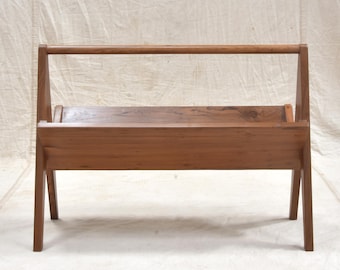 May include: A wooden magazine rack with a handle. The rack is made of light brown wood and has a rectangular shape with angled legs. The handle is a horizontal bar that runs across the top of the rack.