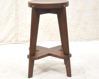 May include: A wooden stool with a round seat and four angled legs. The legs are connected by an X-shaped support near the base. The stool is made of dark brown wood and has a simple, minimalist design.