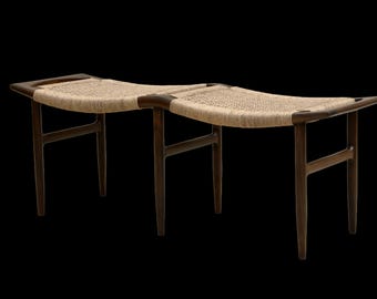 May include: A mid-century modern bench with a woven seat and dark wood frame. The bench has two seating areas and is supported by four angled legs. The woven seat is a light beige color.