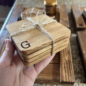 May include: A stack of square wooden coasters tied with twine. Each coaster has a natural wood grain and is engraved with the letter "G". Other wooden items are in the background, including a tray and coasters with the letter "M".
