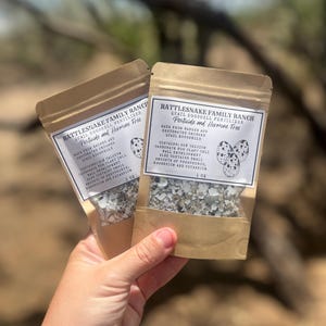 May include: Two small brown paper bags of Rattlesnake Family Ranch quail eggshell fertilizer. The bags have a white label with black text that reads "Pesticide and Hormone Free." The bags are filled with crushed eggshells. The bags are held in a hand.