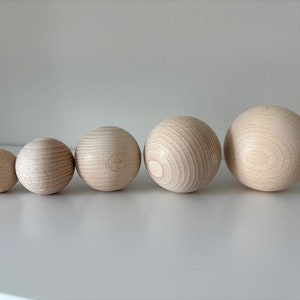 Op de afbeelding: Vijf houten ballen in verschillende maten, gerangschikt in een rij van de kleinste tot de grootste. De ballen zijn van een lichte natuurlijke houtkleur.