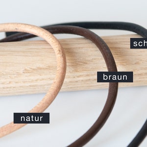 Könnte beinhalten: Drei Lederbänder in verschiedenen Farben: Natur, Braun und Schwarz. Die Armbänder sind auf einer Holzoberfläche abgelegt.