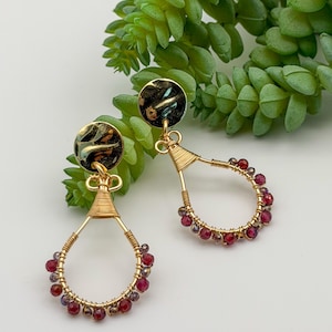 May include: A pair of gold-toned wire-wrapped earrings featuring circular black and gold patterned studs. Each earring has a teardrop-shaped frame adorned with small, faceted deep red and dark purple beads attached with thin gold wire.
