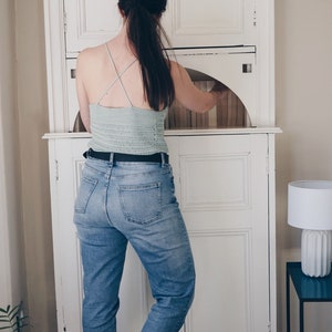 K&ouml;nnte beinhalten: Ein helles, salbeigr&uuml;nes H&auml;kel-Camisole mit d&uuml;nnen, &uuml;berkreuzten Tr&auml;gern am R&uuml;cken, kombiniert mit einer blauen Jeans mit schwarzem G&uuml;rtel. Das Oberteil ist in einem horizontalen Maschenmuster gefertigt und hat eine k&ouml;rpernahe Passform.