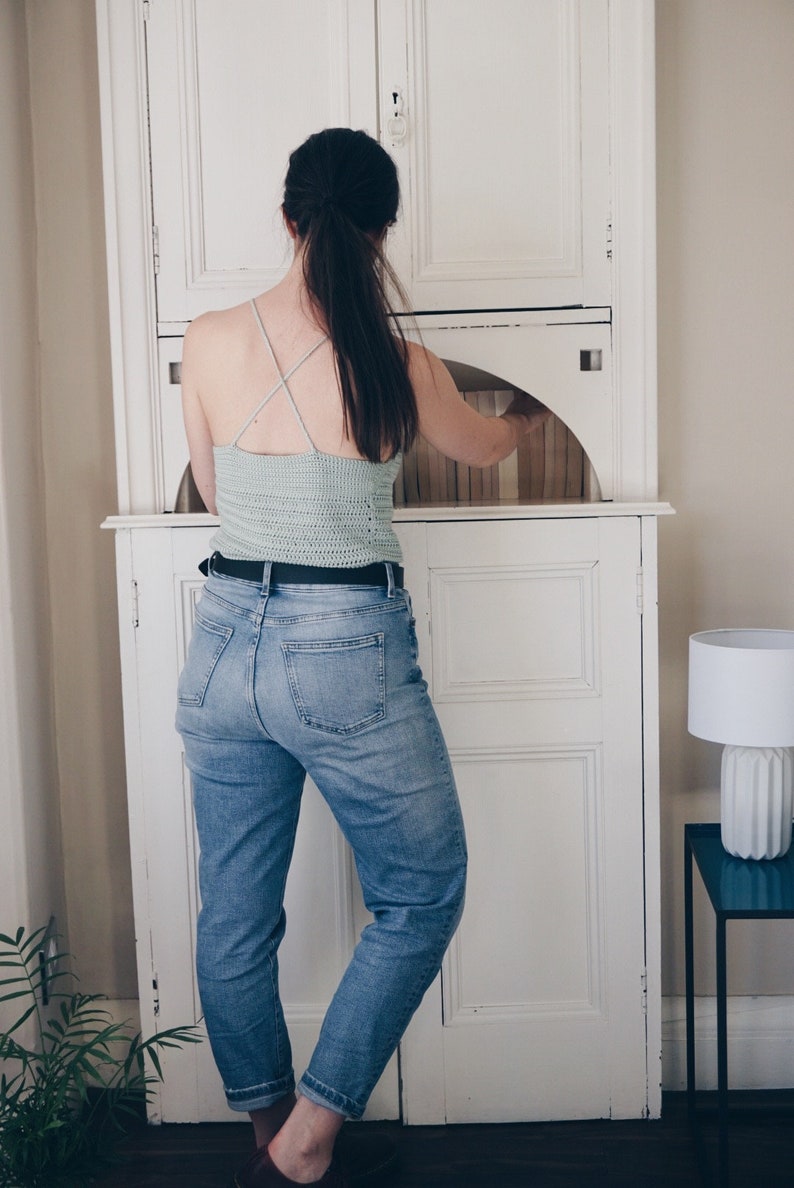 K&ouml;nnte beinhalten: Ein helles, salbeigr&uuml;nes H&auml;kel-Camisole mit d&uuml;nnen, &uuml;berkreuzten Tr&auml;gern am R&uuml;cken, kombiniert mit einer blauen Jeans mit schwarzem G&uuml;rtel. Das Oberteil ist in einem horizontalen Maschenmuster gefertigt und hat eine k&ouml;rpernahe Passform.