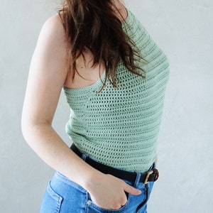 K&ouml;nnte beinhalten: Ein &auml;rmelloses H&auml;keloberteil in hellem Salbeigr&uuml;n mit texturiertem horizontalem Maschenmuster, hohem Ausschnitt und figurbetonter Passform, kombiniert mit einer blauen Jeans und einem schwarzen G&uuml;rtel mit goldfarbener Schnalle.