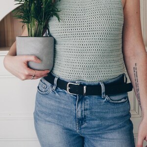 K&ouml;nnte beinhalten: Ein helles, salbeigr&uuml;nes H&auml;kel-Neckholder-Top mit quadratischem Ausschnitt und d&uuml;nnen Tr&auml;gern, kombiniert mit einer blauen Jeans mit schwarzem G&uuml;rtel und silberner Schnalle. Das Top weist ein durchgehendes, horizontales Maschenmuster auf.