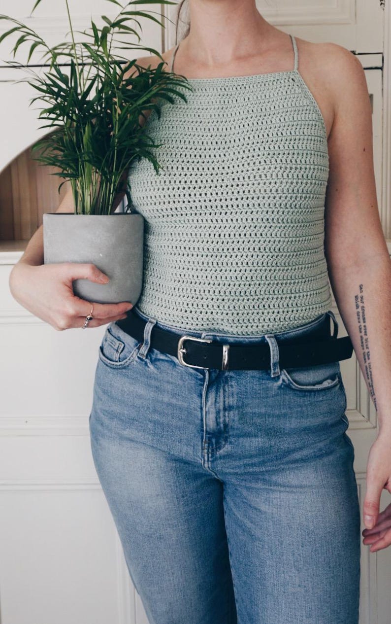 K&ouml;nnte beinhalten: Ein helles, salbeigr&uuml;nes H&auml;kel-Neckholder-Top mit quadratischem Ausschnitt und d&uuml;nnen Tr&auml;gern, kombiniert mit einer blauen Jeans mit schwarzem G&uuml;rtel und silberner Schnalle. Das Top weist ein durchgehendes, horizontales Maschenmuster auf.