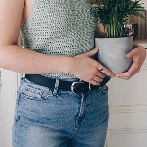K&ouml;nnte beinhalten: Ein helles, salbeigr&uuml;nes H&auml;kel-Neckholder-Top mit d&uuml;nnen Tr&auml;gern, kombiniert mit einer blauen Jeans und einem schwarzen Lederg&uuml;rtel mit silberner Schnalle. Das Top hat ein strukturiertes Webmuster und einen hohen Ausschnitt f&uuml;r eine Passform.
