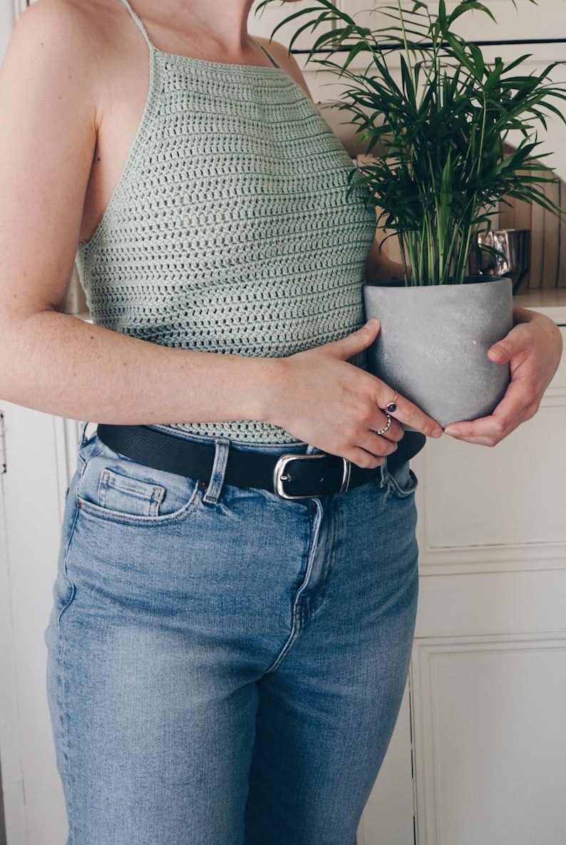 K&ouml;nnte beinhalten: Ein helles, salbeigr&uuml;nes H&auml;kel-Neckholder-Top mit d&uuml;nnen Tr&auml;gern, kombiniert mit einer blauen Jeans und einem schwarzen Lederg&uuml;rtel mit silberner Schnalle. Das Top hat ein strukturiertes Webmuster und einen hohen Ausschnitt f&uuml;r eine Passform.