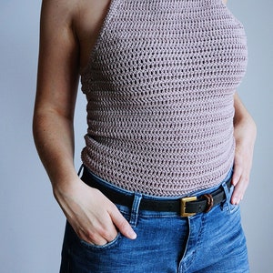 K&ouml;nnte beinhalten: Ein &auml;rmelloses H&auml;kel-Neckholder-Top in Mauve mit quadratischem Ausschnitt und d&uuml;nnen Tr&auml;gern, kombiniert mit einer blauen Jeans, die mit einem schwarzen Lederg&uuml;rtel mit messingfarbener Schnalle gehalten wird. Das Top hat ein strukturiertes Webmuster.