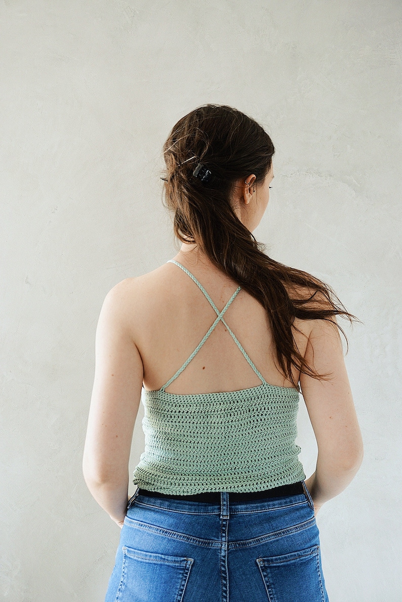 K&ouml;nnte beinhalten: Ein helles, salbeigr&uuml;nes H&auml;kel-Camisole mit geradem Ausschnitt und d&uuml;nnen, geflochtenen Spaghettitr&auml;gern, die sich am R&uuml;cken kreuzen. Das Kleidungsst&uuml;ck ist mit einem horizontalen Maschenmuster gefertigt und wird mit einer blauen Jeans kombiniert.