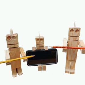 May include: Three wooden robot figures holding pencils, with one supporting a black smartphone. The robots are made of natural wood and have simple, blocky designs. One pencil has the text "THE HOME DEPOT".