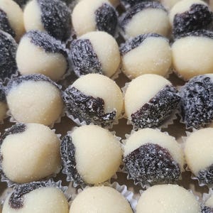 May include: A collection of round, bite-sized confections arranged in a grid. Each piece features a dual-tone appearance with a smooth, creamy white section and a textured, dark brown section, all lightly dusted with fine sugar crystals.
