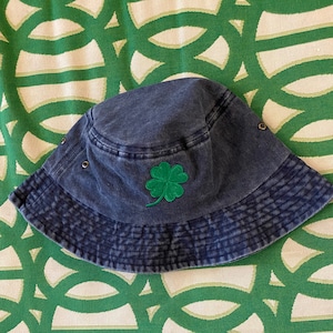May include: A denim blue bucket hat with a green four-leaf clover patch. The hat has a faded, worn look and features a structured brim. The hat is a casual accessory.