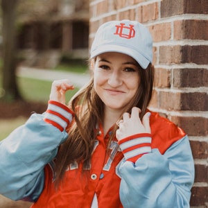 May include: A light blue baseball cap with a red embroidered "UD" logo. The person is wearing a red and light blue satin varsity jacket with white and red striped cuffs. The jacket has a button closure.