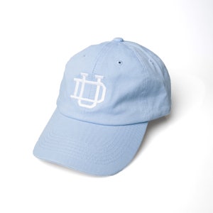 May include: A light blue baseball cap with a curved brim. The cap features a white embroidered logo on the front. The hat is made of a soft fabric and has a classic six-panel design. The cap is a solid light blue color.