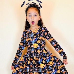 May include: A long-sleeved black dress with a Halloween-themed print featuring jack-o'-lanterns and witches. The dress is worn with a bat-shaped headband. The dress has a tiered skirt and a round neckline. The girl is looking at the camera with an open mouth.