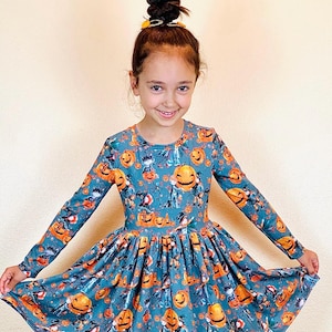 May include: A long-sleeved teal dress featuring a Halloween-themed print. The dress is adorned with orange jack-o'-lanterns, bats, and other spooky characters. The dress has a round neckline and a flared skirt, perfect for a festive occasion.