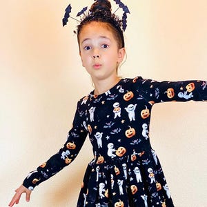 May include: A black Halloween dress with long sleeves, featuring a pattern of pumpkins, ghosts, bats, and stars. The dress has a round neckline and a flared skirt. A black headband with bat decorations completes the festive look.