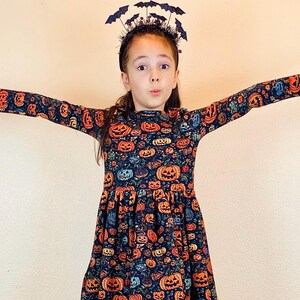 May include: A long-sleeved dress with a pumpkin pattern in orange, blue, and green. The dress is paired with a black headband adorned with bat silhouettes, perfect for Halloween. The dress has a gathered skirt.