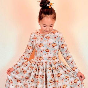 May include: A long-sleeved dress featuring a light blue background with a repeating pattern of orange pumpkins and floral accents. The dress has a round neckline and a full skirt, suitable for fall or autumn-themed events.