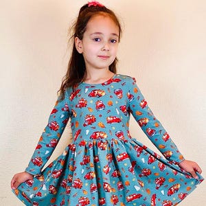 May include: A long-sleeved, teal dress featuring a pattern of red fire trucks, basketballs, and autumn leaves. The dress has a round neckline and a flared skirt. The girl is wearing a pink hair accessory.
