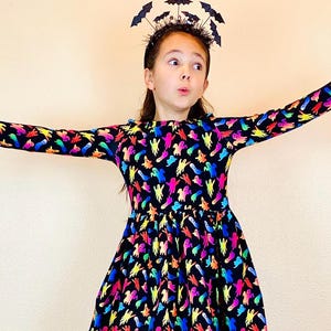 May include: A black dress with long sleeves and a colorful ghost pattern. The dress has a round neckline and a gathered waist. A black headband with bat decorations is worn on the head. The dress is suitable for Halloween.