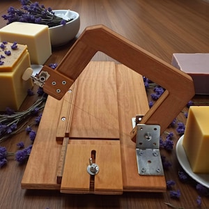 May include: A wooden soap cutter with a handle, string, and metal hardware. Several bars of soap in shades of yellow and purple are arranged around the cutter, along with sprigs of purple flowers. The cutter is on a wooden surface.