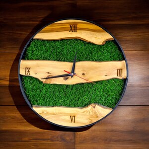 May include: A round wooden clock with Roman numerals and a black clock hand. The clock face is surrounded by a black frame and features a green grass-like material.