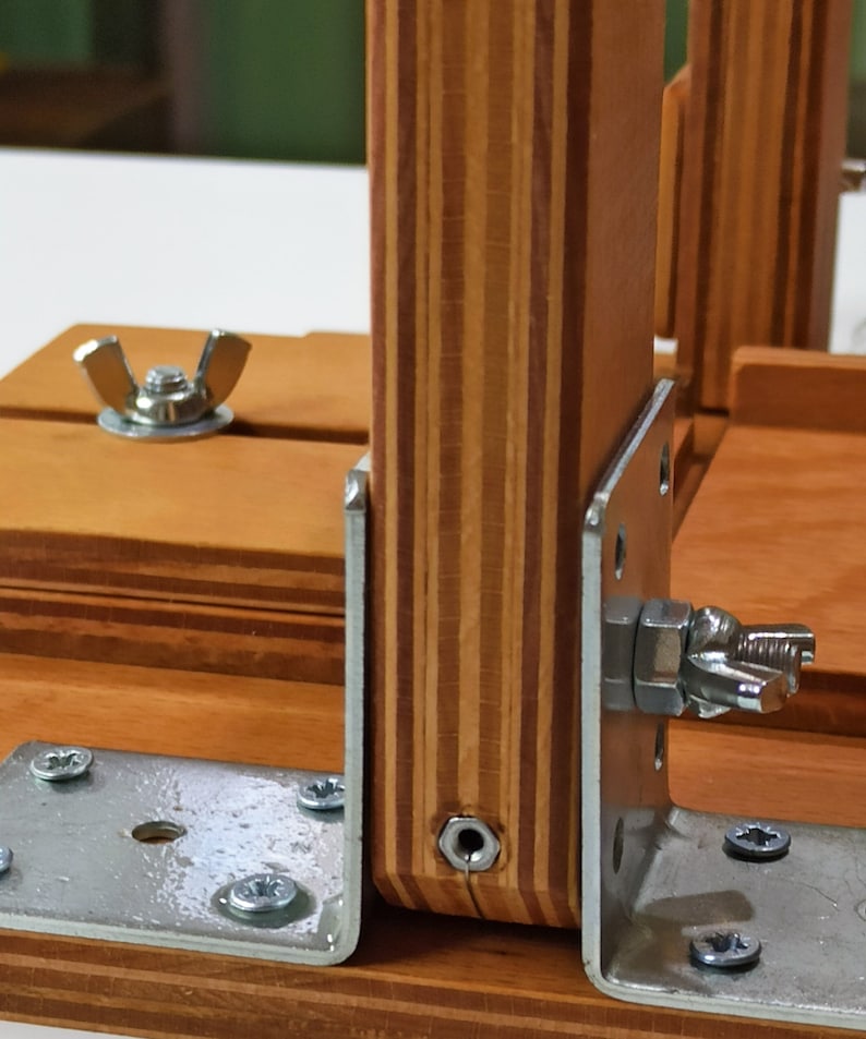Pu&ograve; includere: Primo piano di una cornice in legno con staffe metalliche e dadi a farfalla. La cornice &egrave; realizzata in compensato con un motivo a strisce. Le staffe sono fissate alla cornice con viti.