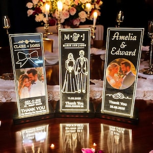 May include: Three illuminated wedding table decorations. Each features a couple's name, date, and a photo or illustration. The decorations are black with white text and a warm, glowing light. The bases are dark.
