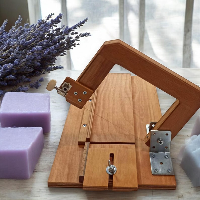 Pu&ograve; includere: Uno strumento di taglio per sapone in legno con un filo e una manopola di tensione. Lo strumento viene utilizzato per tagliare le barre di sapone in pezzi pi&ugrave; piccoli. Nell'immagine ci sono tre barre di sapone viola.